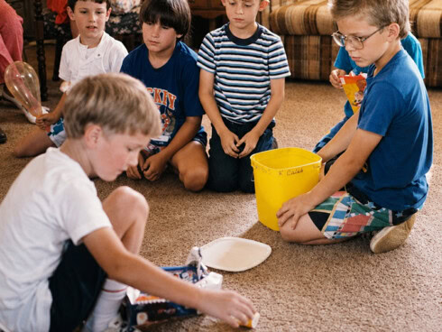 A group of young boys sits on a living room floor during a childhood gathering. In the foreground, a boy in a white shirt plays with a toy, while another boy in a blue shirt and glasses kneels beside a bright yellow bucket.